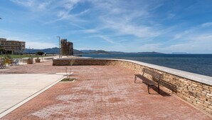 Un paseo marítimo con muro de piedra, un banco y vistas al mar y montañas distantes.