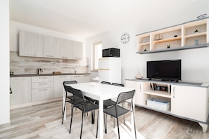 Una cocina moderna con armarios blancos, una mesa de comedor con sillas, un microondas, un televisor sobre un mueble bajo y un reloj en la pared.