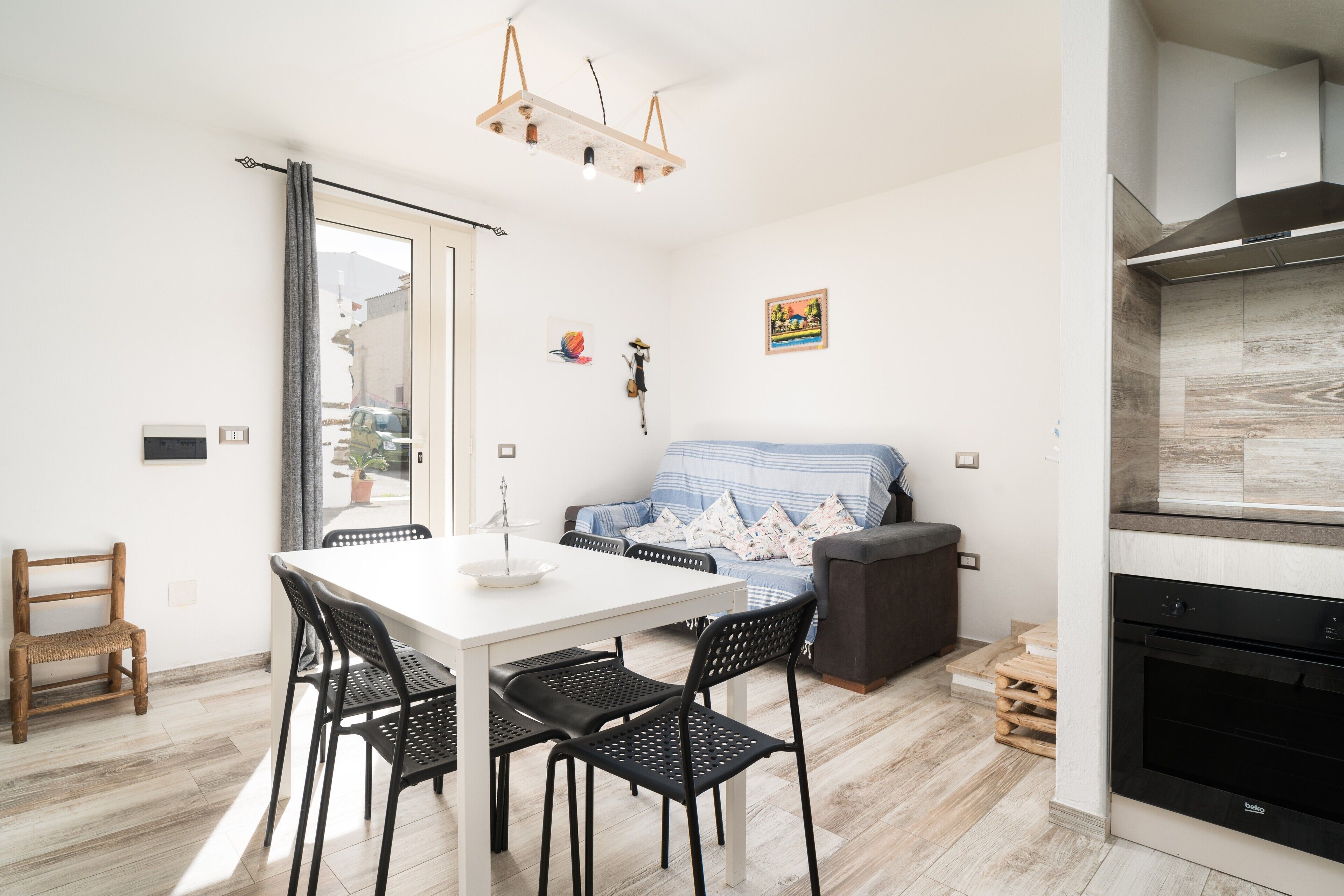 Une cuisine moderne dotée d’un four encastré, un coin repas avec une table blanche et des chaises noires, et un espace de détente avec un canapé et des coussins.