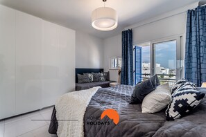 A modern bedroom with a large bed, a bedside table, a wardrobe, and a view of the city through a large window.