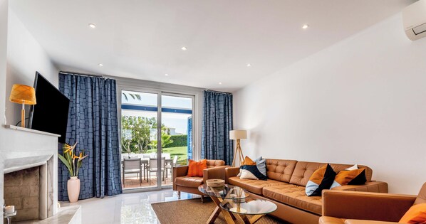 A modern living room with a sofa set, a fireplace, a flat-screen TV, and a large sliding glass door leading to an outdoor area.