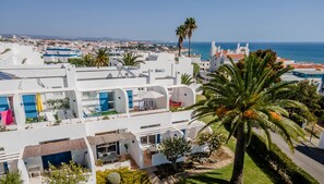 A white building with multiple balconies, a palm tree, and a clear view of the sea.