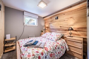 Een slaapkamer met een houten hoofdeinde, een bed met bloemenbeddengoed, een nachtkastje met een lamp en een raam met uitzicht op een gebouw.