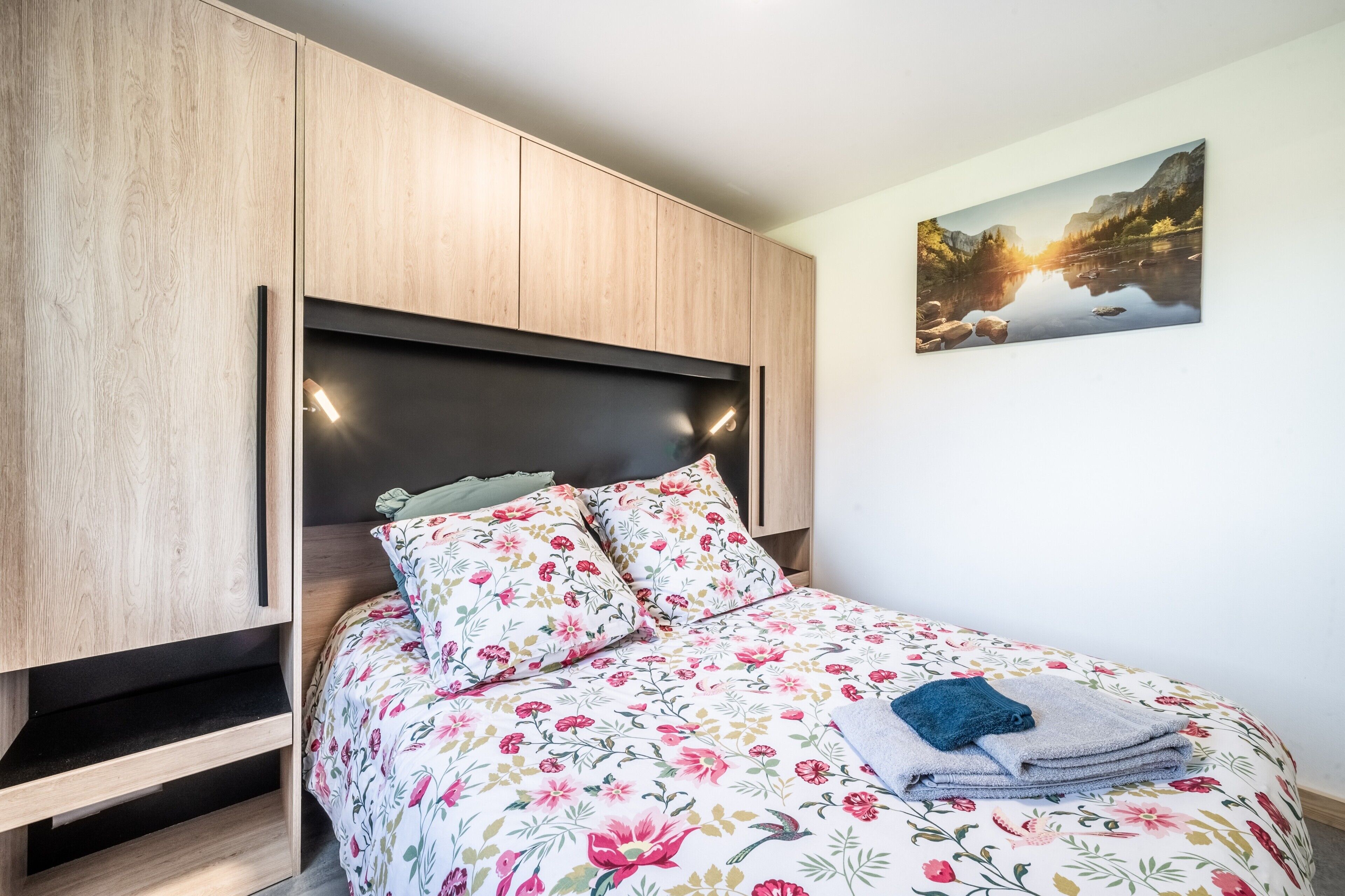 A bedroom with a bed, floral bedding, a nightstand, a wooden wardrobe, and a landscape painting on the wall.
