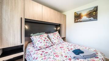 A bedroom with a bed, floral bedding, a nightstand, a wooden wardrobe, and a landscape painting on the wall.
