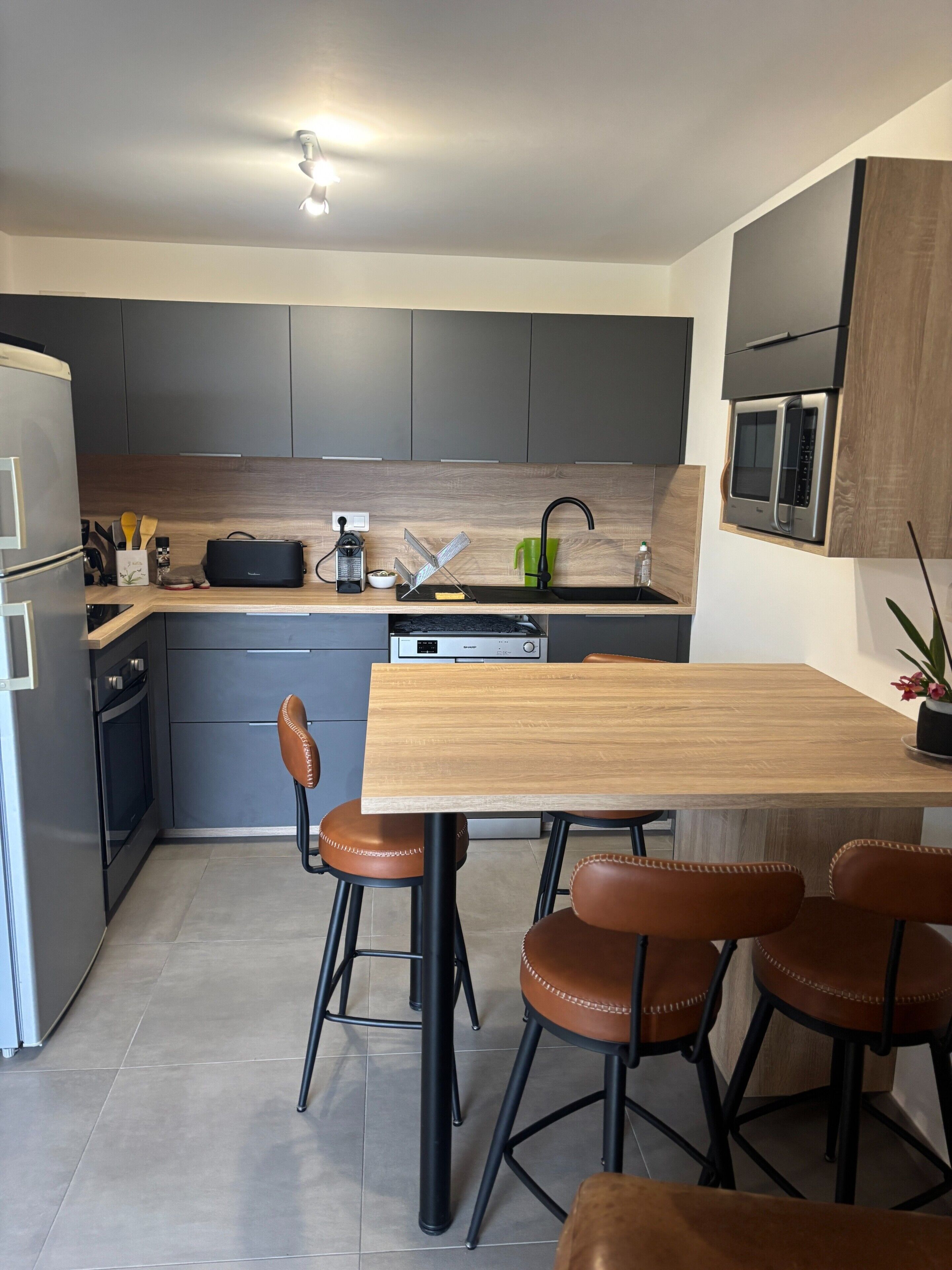 A modern kitchen with grey cabinets, a wooden countertop, a built-in microwave, and a dining area with a wooden table and leather stools.