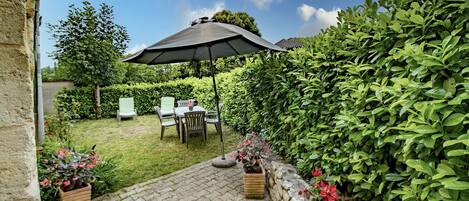 Un patio con mesa y sillas bajo un gran paraguas, rodeado de exuberante vegetación y un muro de piedra.