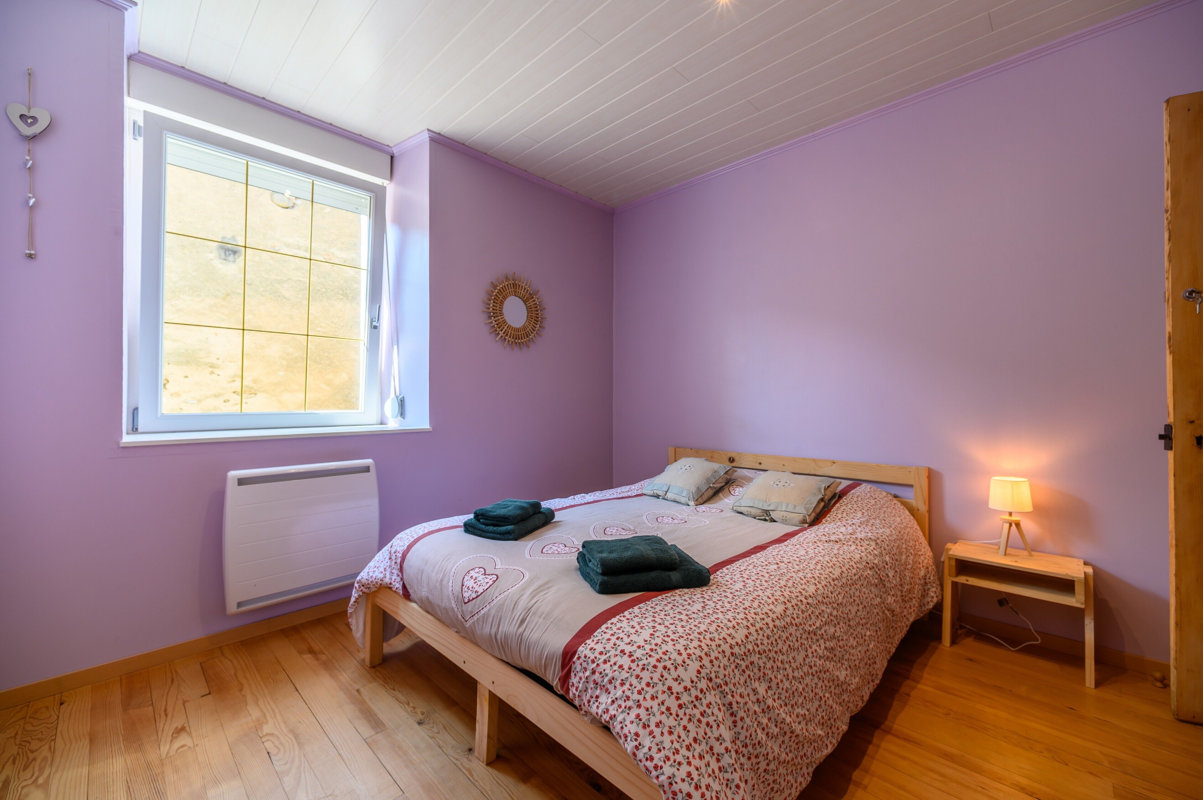 Een slaapkamer met een bed, een nachtkastje met een lamp, een raam met zonwering, een radiator en een wanddecoratie.