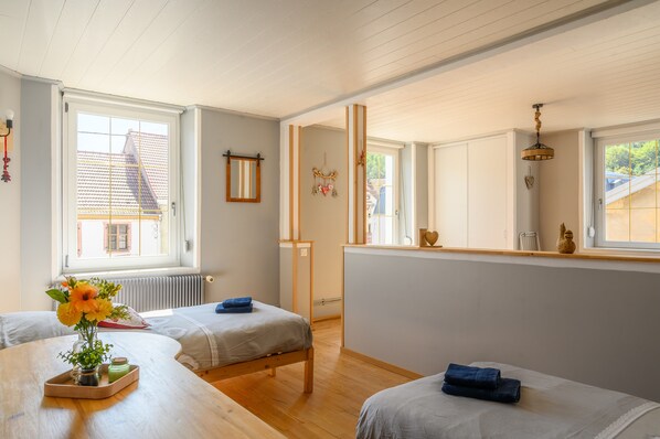 A room with two beds, a wooden table with a vase, a window with a view, and a mirror on the wall.