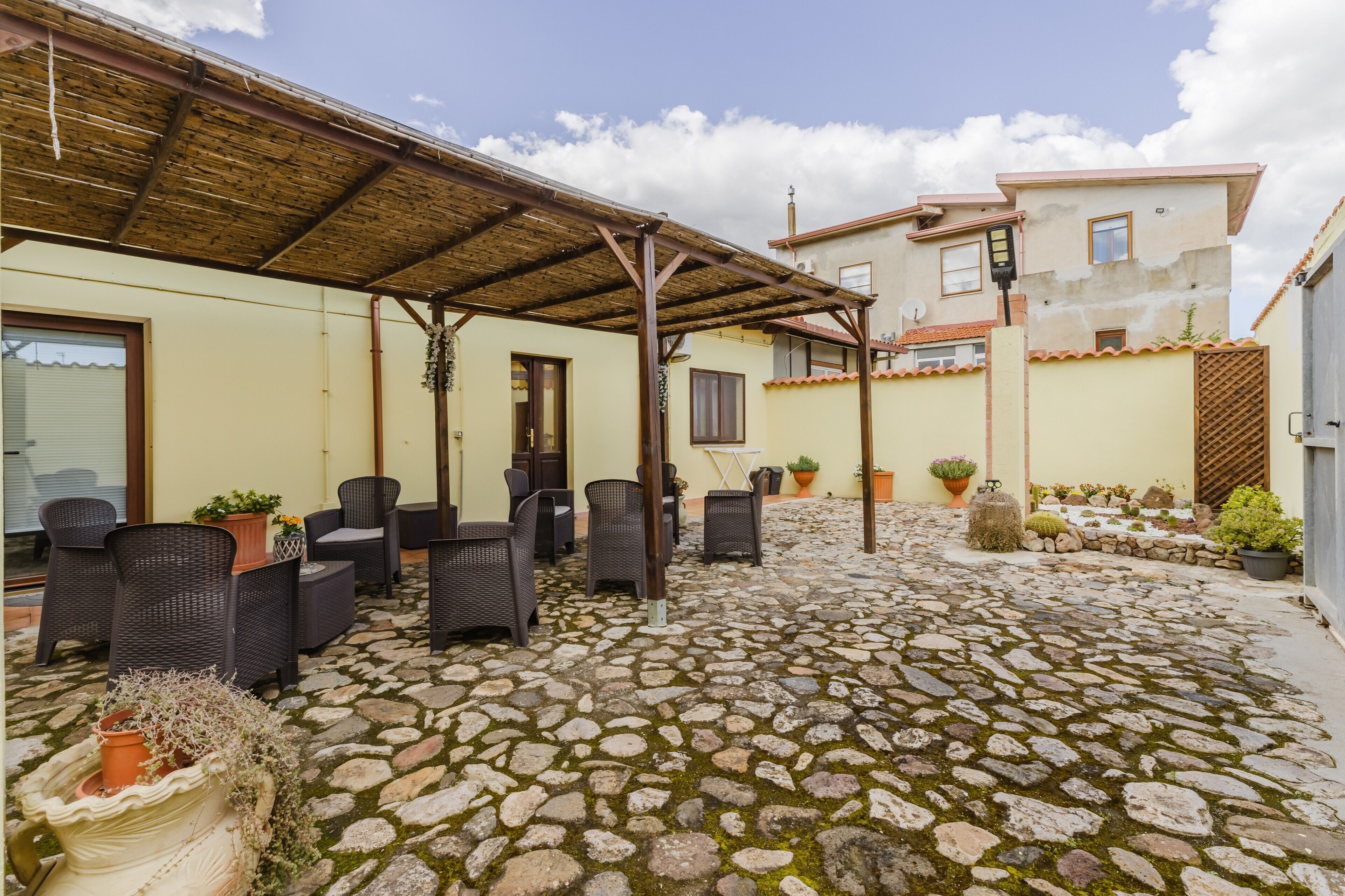 A covered outdoor seating area with wicker furniture, a stone-paved patio, and a building with a tiled roof in the background.