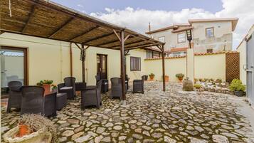A covered outdoor seating area with wicker furniture, a stone-paved patio, and a building with a tiled roof in the background.