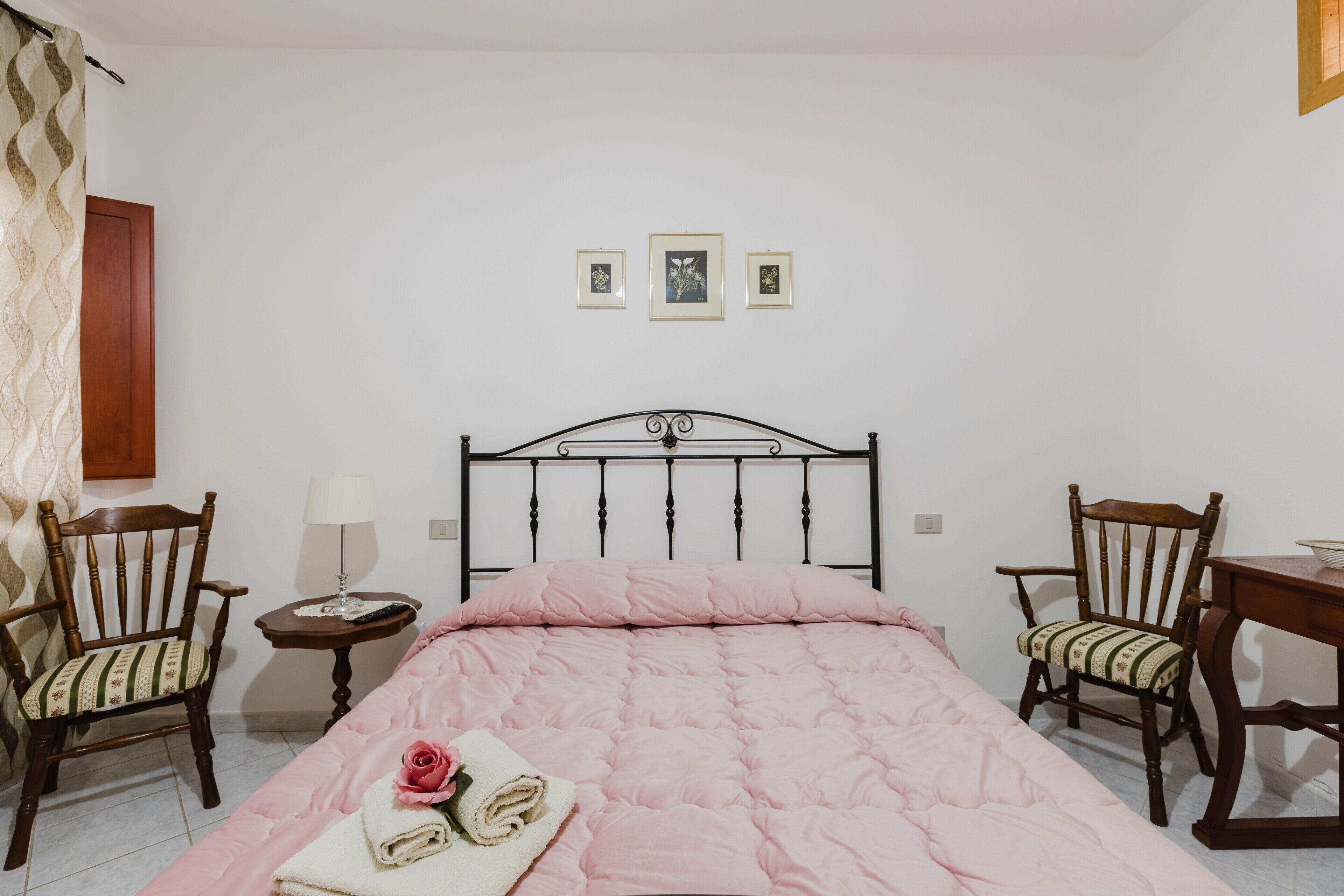 A bedroom with a bed, two chairs, a small table, and three framed pictures on the wall.