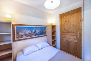 A bedroom with a bed, a wooden door, a shelf with a lamp, and a framed picture on the wall.