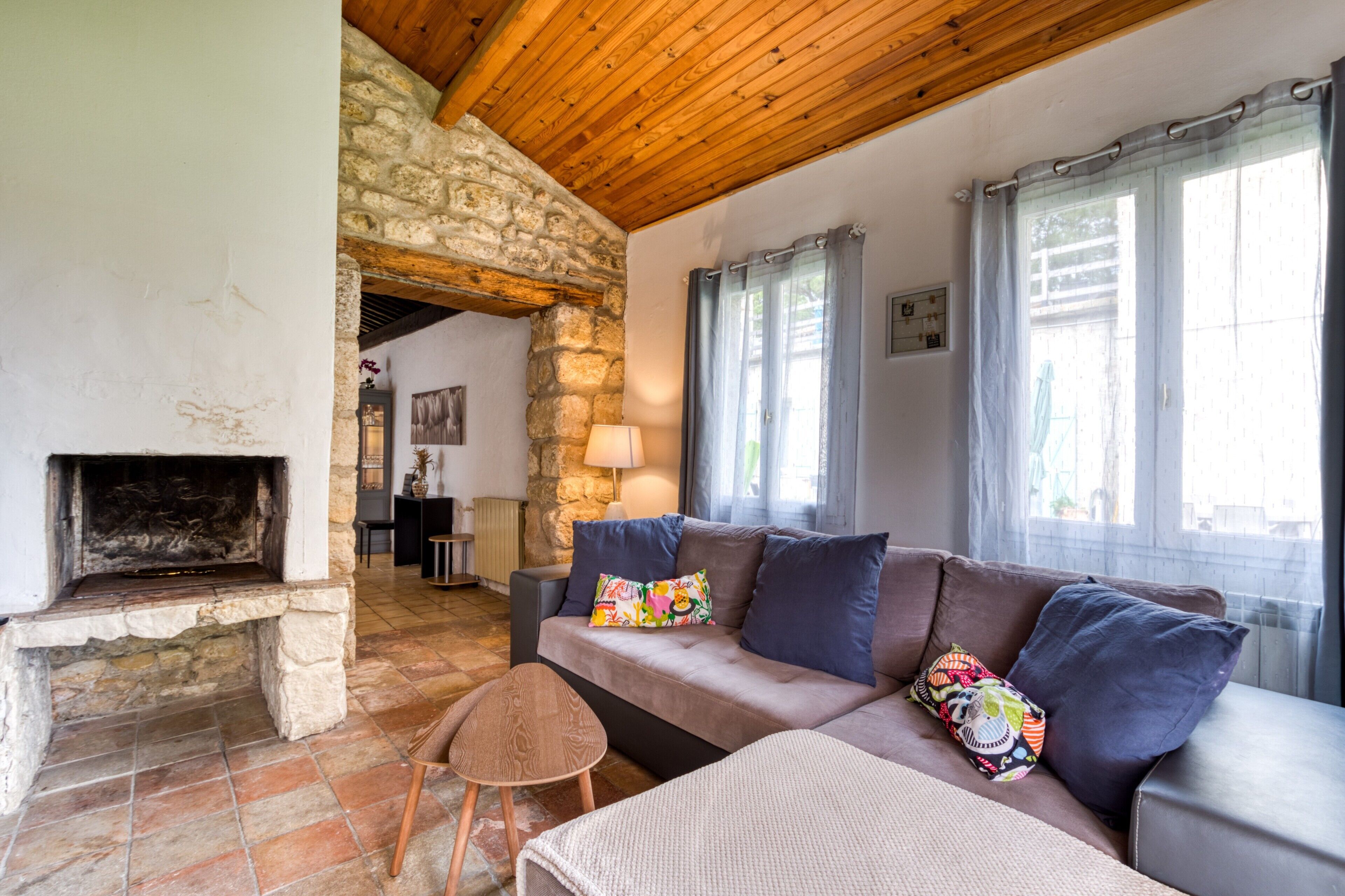 A cozy living room with a stone fireplace, a wooden ceiling, and a sofa with colorful cushions.