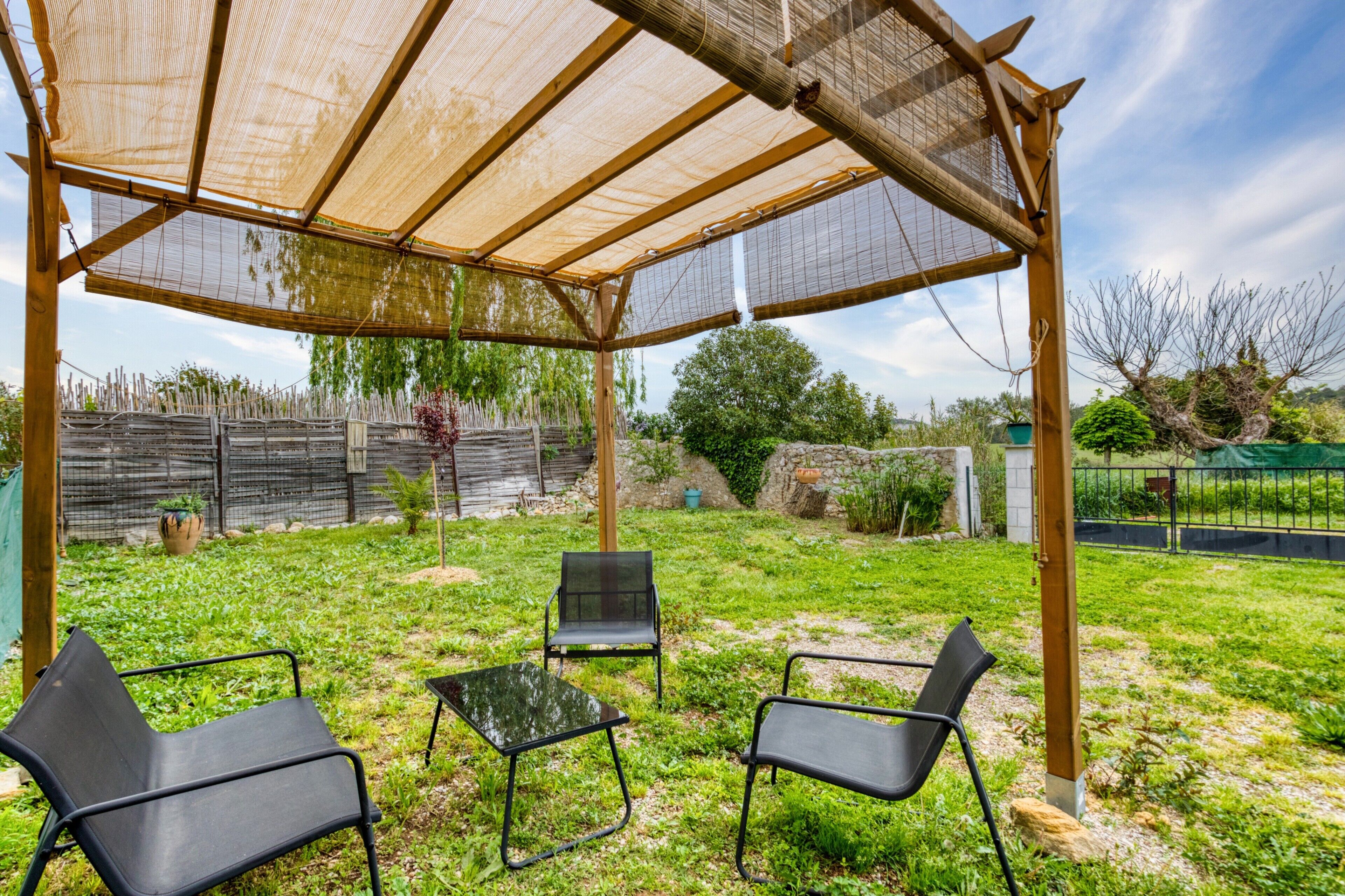 An outdoor covered patio with three chairs and a table, surrounded by a grassy area and a wooden fence.