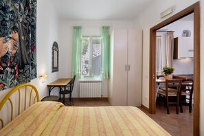 A bedroom with a bed, a window with curtains, a radiator, a desk with a chair, and a dining area with chairs and a table.