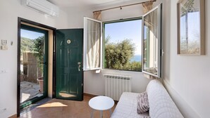Una habitación con cama, una mesita, un radiador, una ventana con cortinas y una puerta abierta que da a un espacio exterior.