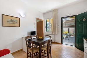 A dining area with a table, chairs, a basket of fruit, a painting on the wall, a computer, and a view through a door to an outdoor patio.