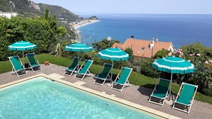 A pool with clear water, surrounded by green lounge chairs and umbrellas, with a view of the sea and a mountainous landscape. - Apartment 'La Realidad 3' with Shared Pool, Wi-Fi and Air Conditioning (Finale Ligure)