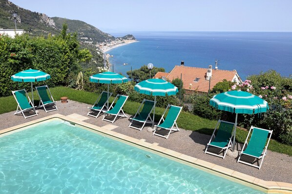 A pool with clear water, surrounded by green lounge chairs and umbrellas, with a view of the sea and a mountainous landscape.