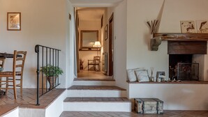 Una escalera conduce a un comedor con sillas y una mesa, una chimenea con adornos, y una planta en la barandilla.