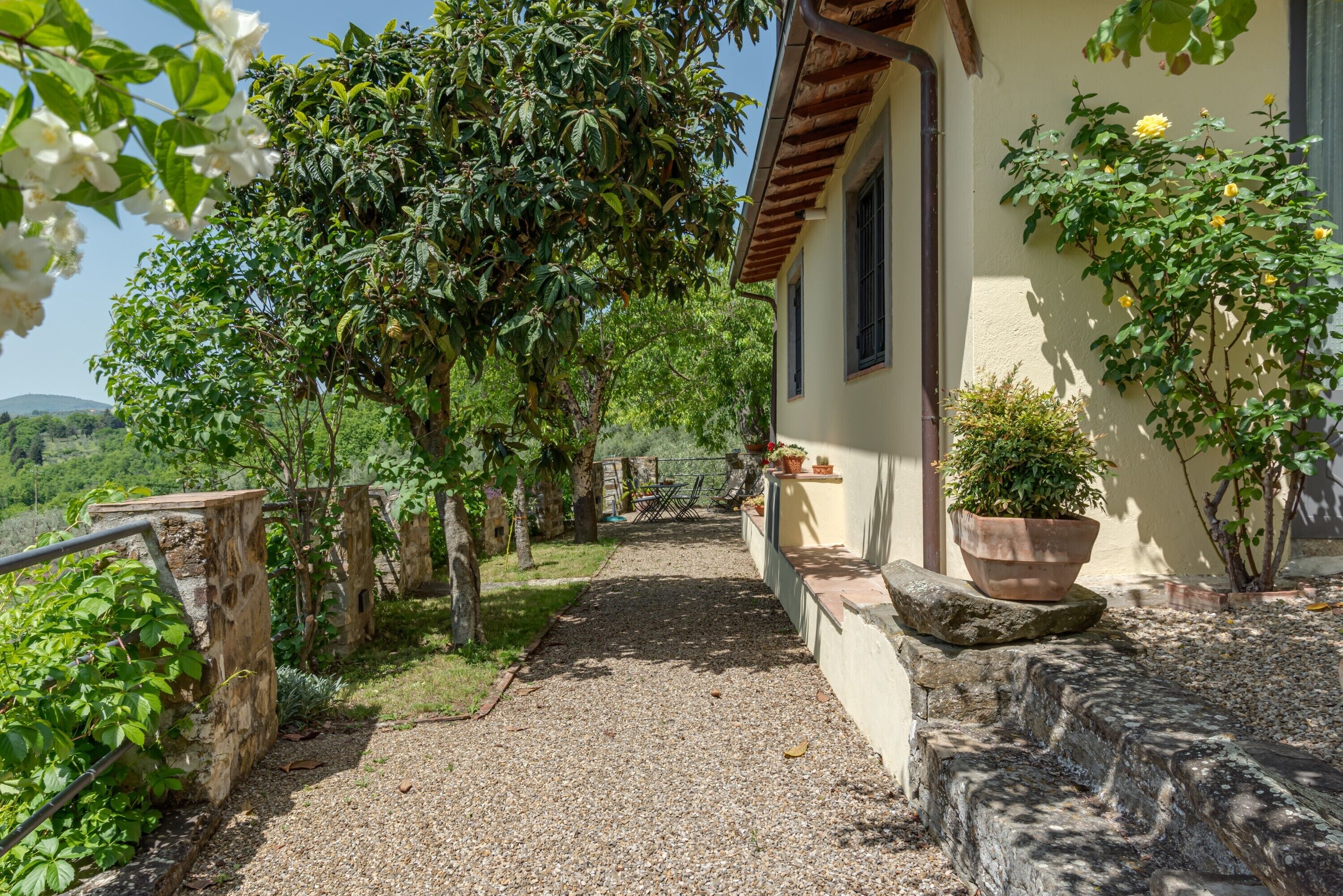 A pathway leading to a building with a stone wall, potted plants, and a view of the countryside.
