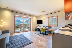 A modern living room with a sofa, a dining area with chairs, a kitchen area, and large windows with a mountain view.