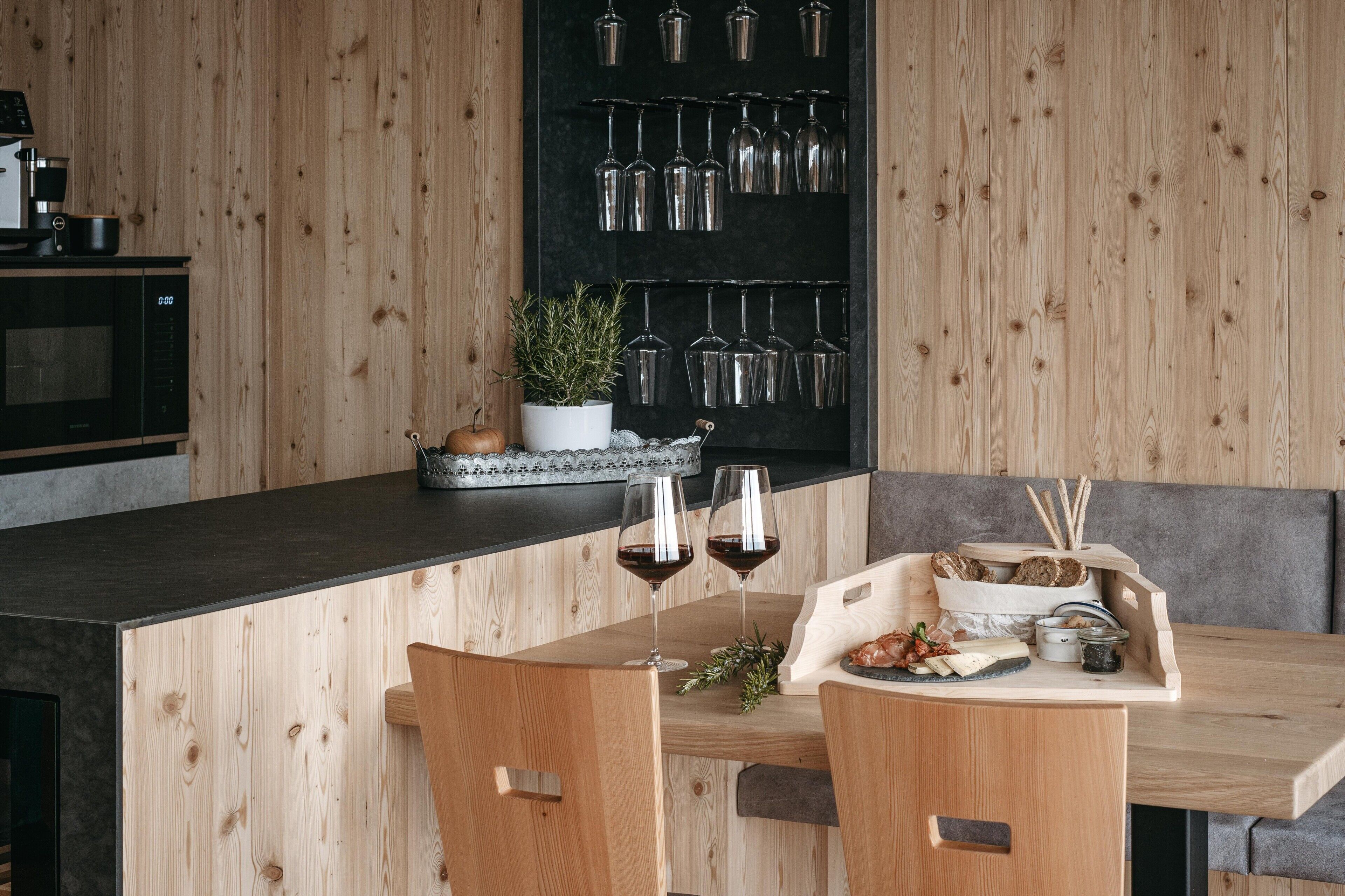 Een houten eethoek met een zwart aanrecht, twee stoelen, een kleine tafel met wijnglazen, een potplant en een wandmontage wijnrek.