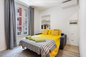 Une chambre d’hôtel avec un lit, une fenêtre avec des rideaux, un climatiseur fixé au mur et une petite étagère avec des livres et un tableau.