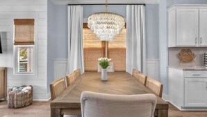 Ein Esszimmer mit Holztisch, weißen Vorhängen, einer Chandelier und einer Küchenzeile mit weißen Schränken.