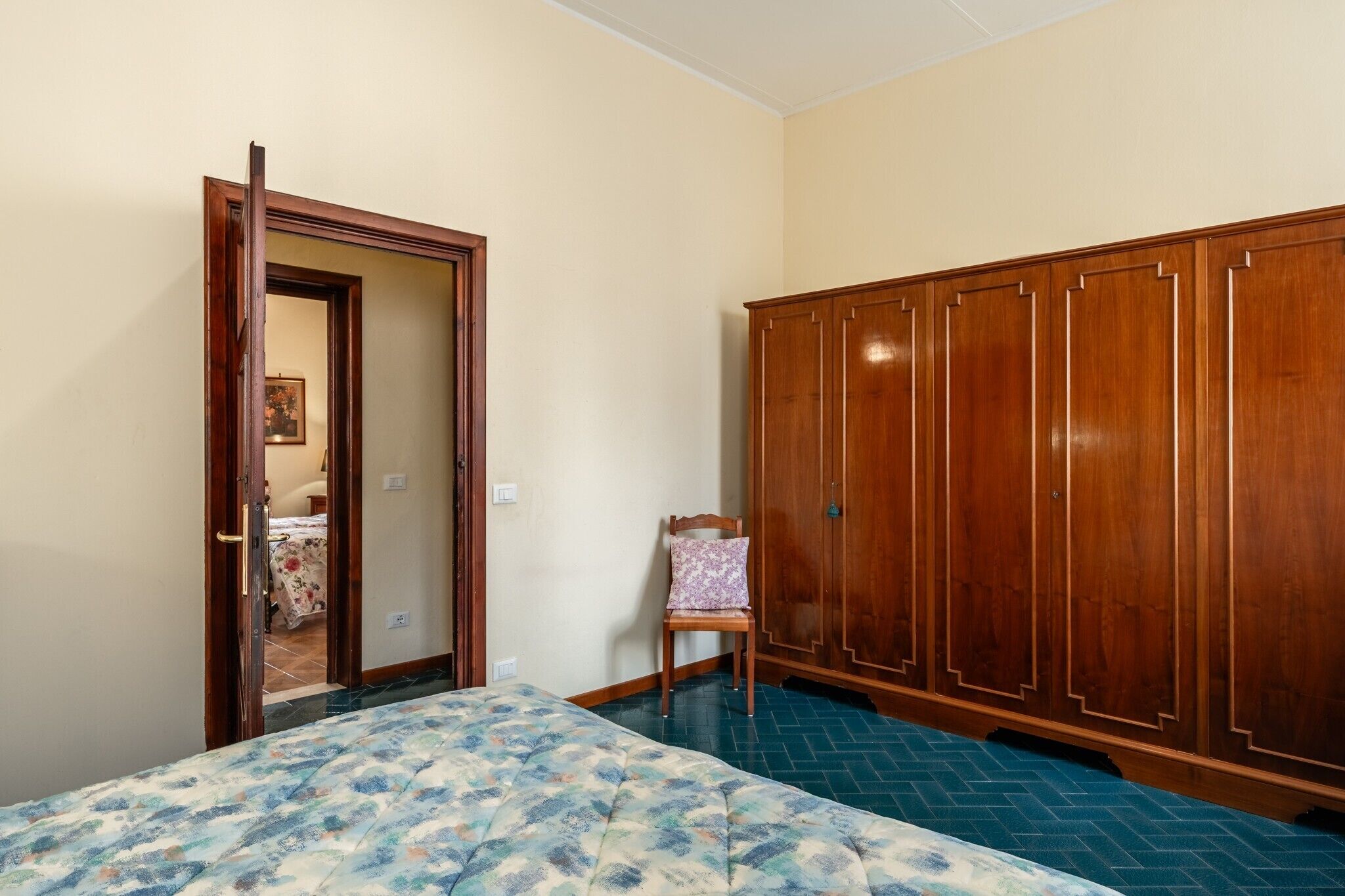 A bedroom with a bed, a wooden wardrobe, a chair, and a door leading to another room.