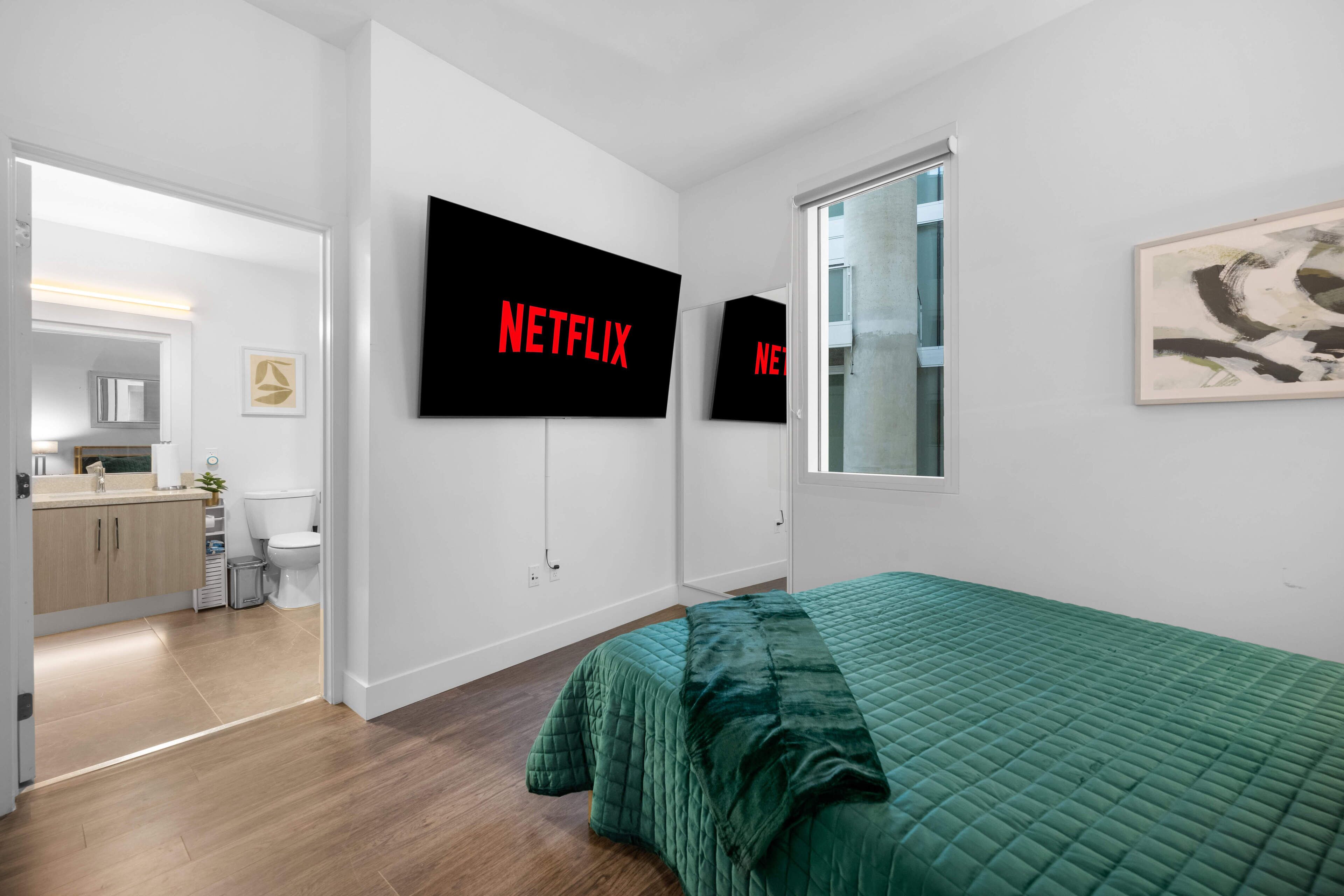 A modern bedroom with a green bedspread, a large flat-screen TV mounted on the wall, and a view of the bathroom through an open door.