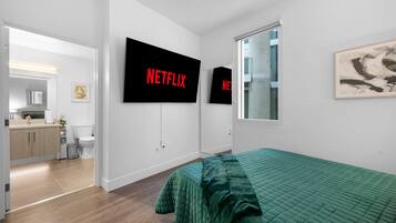 A modern bedroom with a green bedspread, a large flat-screen TV mounted on the wall, and a view of the bathroom through an open door.