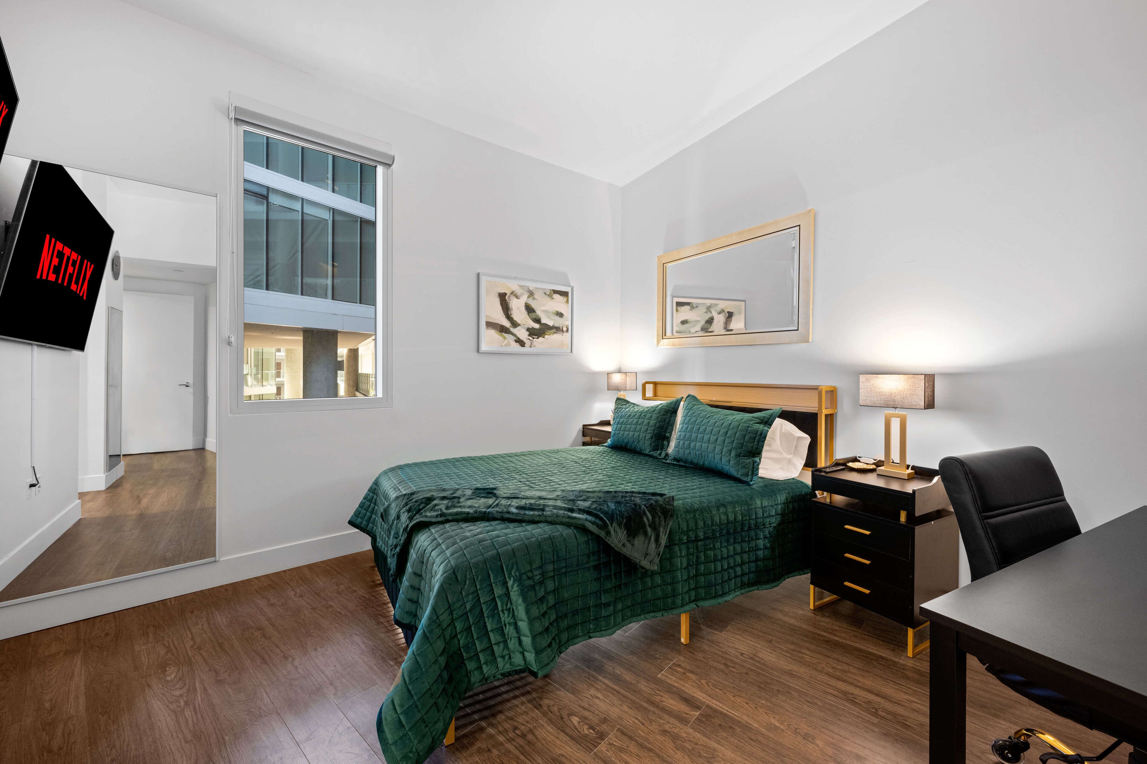 Un dormitorio moderno con una cama verde, suelo de madera, un escritorio, una silla, un televisor y obras de arte en las paredes.