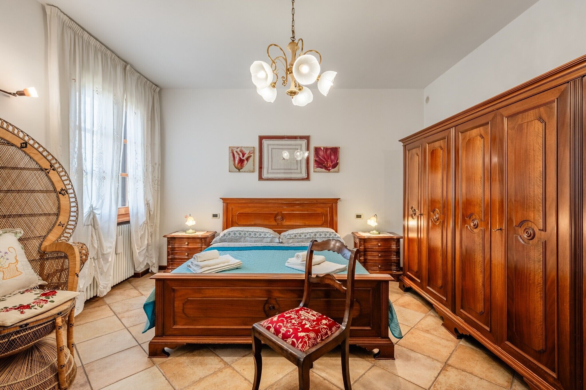 A bedroom with a wooden bed, a wooden wardrobe, a wicker chair, a red upholstered chair, and a chandelier.