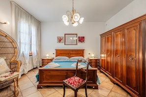 Une chambre à coucher avec un lit en bois, une armoire en bois, une chaise en osier, un fauteuil recouvert de tissu rouge et un lustre.
