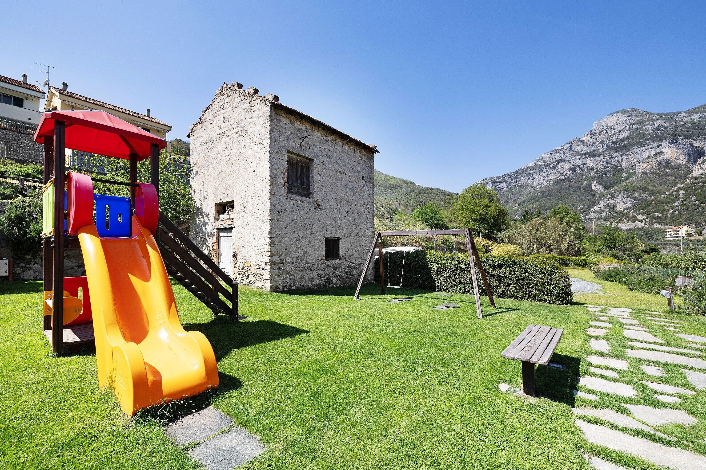 Ein Spielplatz mit Rutsche, Picknicktisch und einem Steinhaus in einer grünen Umgebung, im Hintergrund Berge.