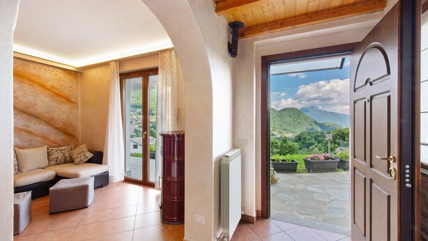 Een kamer met een houten plafond, een bank, een radiator en een deur naar een balkon met uitzicht op de bergen.