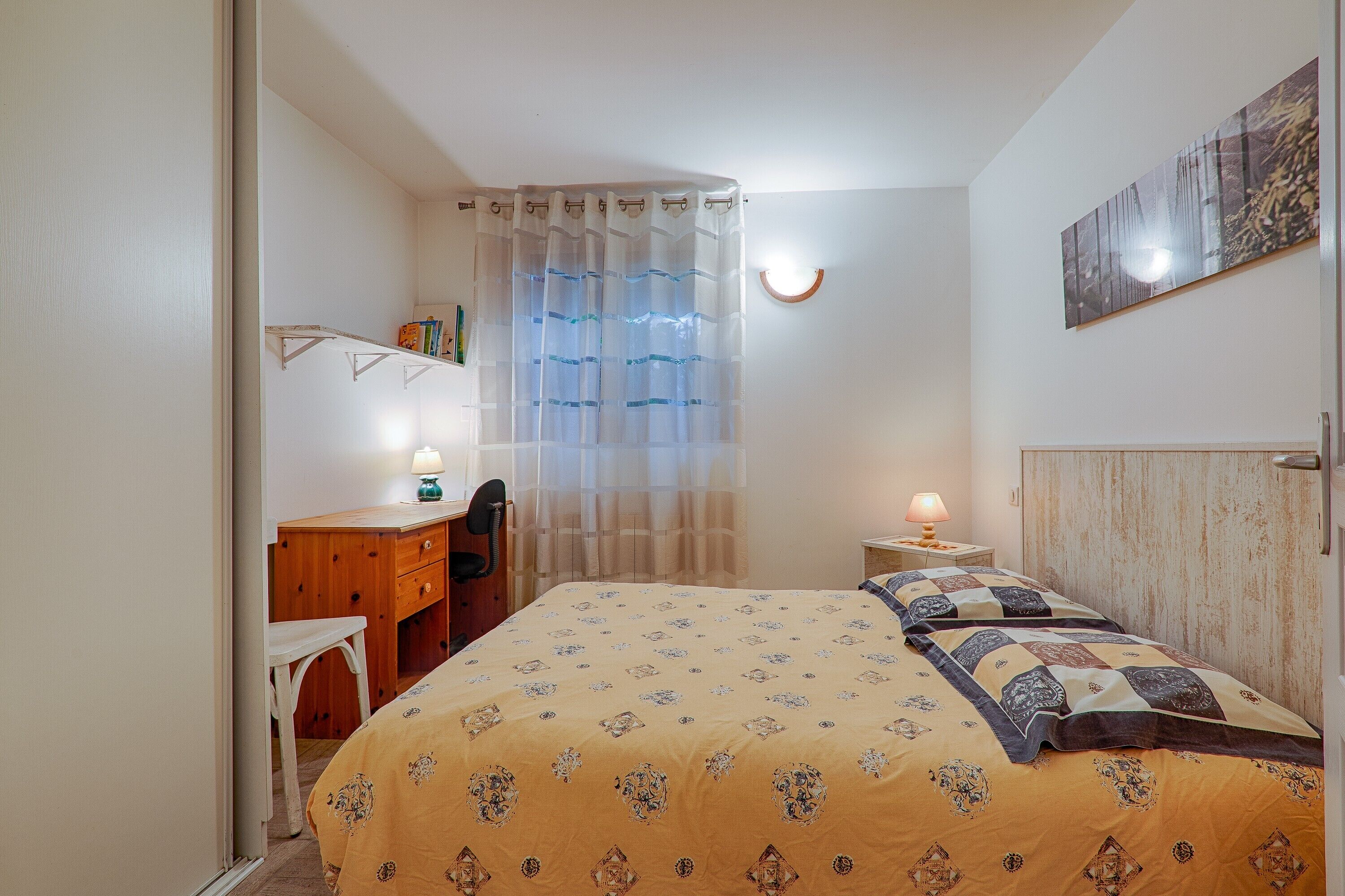A bedroom with a bed, a desk, a chair, a nightstand with a lamp, and a window with curtains.