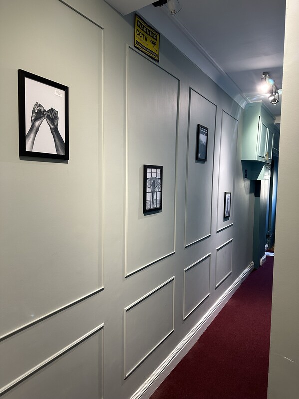 A hallway with framed artwork on the walls, a CCTV warning sign, and a door leading to a room with teal cabinets.