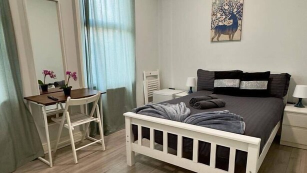 A bedroom with a bed, a desk, a chair, a nightstand, a lamp, a painting, and a window with curtains. - Guest&Rest Hotel (London)