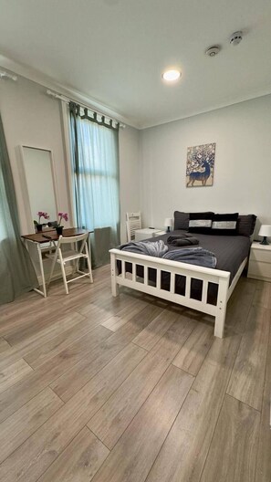 A bedroom with a bed, a desk, a chair, a nightstand, a lamp, a painting, and a window with curtains.