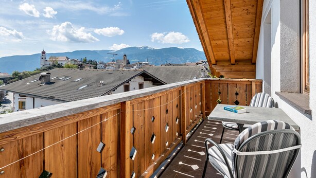 Un balcón con vistas a edificios y montañas, barandas de madera y una mesa con sillas.