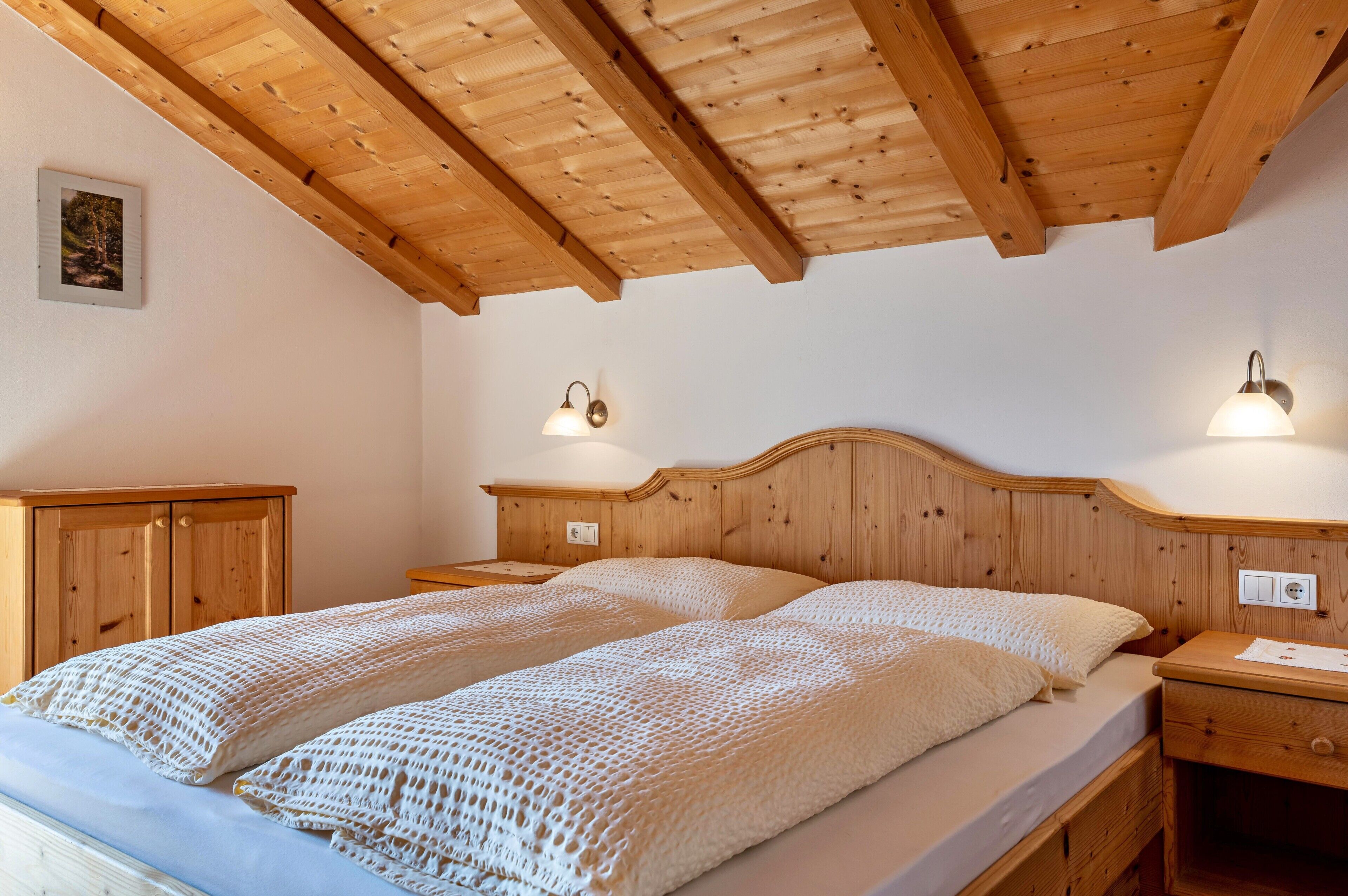A wooden ceiling, a bed with a headboard, two bedside tables, a nightstand, and a wall-mounted light fixture.