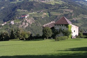 Um edifício branco com telhado vermelho, rodeado por um campo verde e montanhas ao fundo.