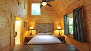 A wooden cabin bedroom with a bed, two bedside lamps, a ceiling fan, and a window with curtains. - Great small cabin with hot tub (Bryson City)