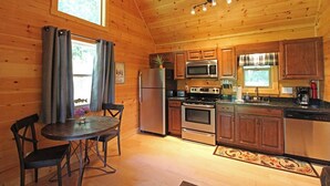 A wooden cabin interior with a kitchen featuring a refrigerator, microwave, stove, and sink. There is a dining table with chairs and a window with curtains. - Great small cabin with hot tub (Bryson City)