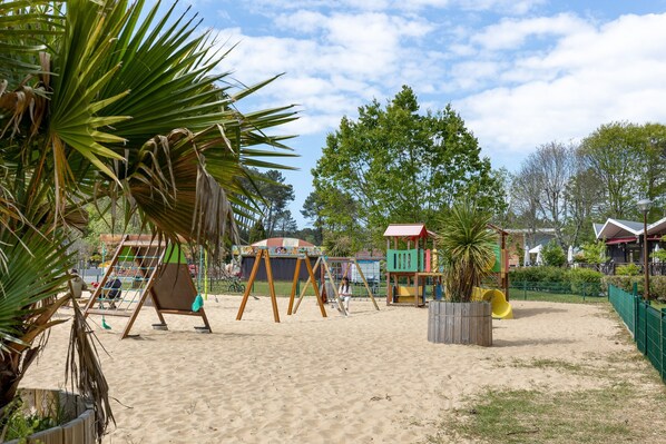 Un parque infantil de arena con columpios y un tobogán, rodeado de árboles y edificios.