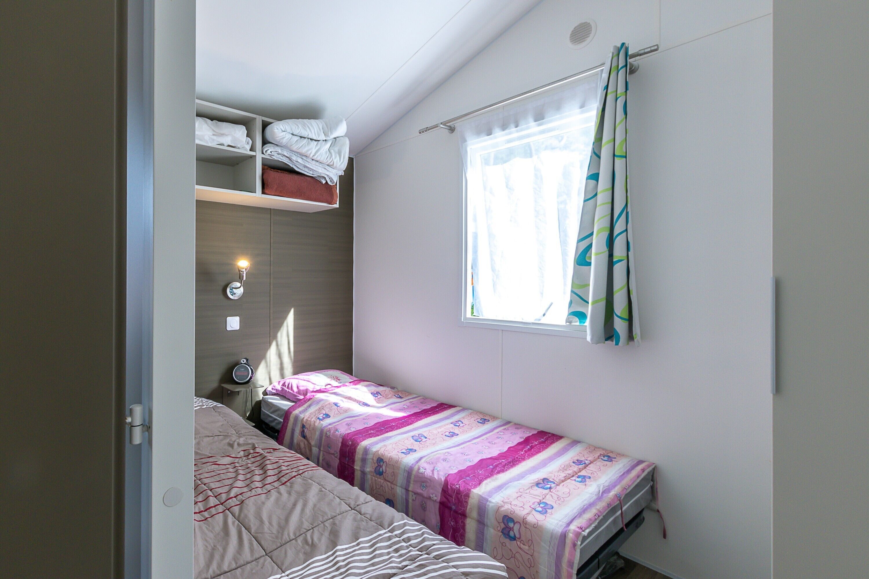 A small bedroom with a single bed, a nightstand, a shelf with folded towels, and a window with curtains.