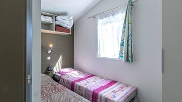 Un dormitorio pequeño con una cama individual, una mesita de noche, un estante con toallas dobladas y una ventana con cortinas.